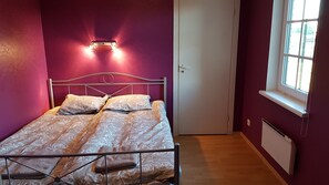 A bedroom with a bed, a nightstand, a wall-mounted light, and a window. - Sinilille 7 Holiday Home (Kuressaare)