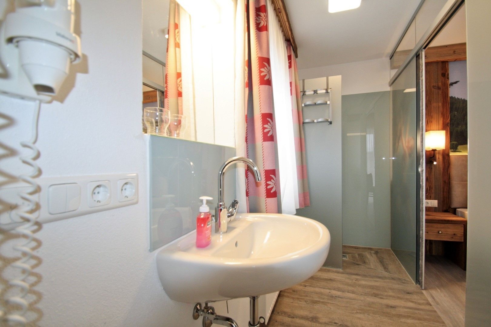 Un baño con lavamanos blanco, un espejo, zona de ducha y suelo de madera.