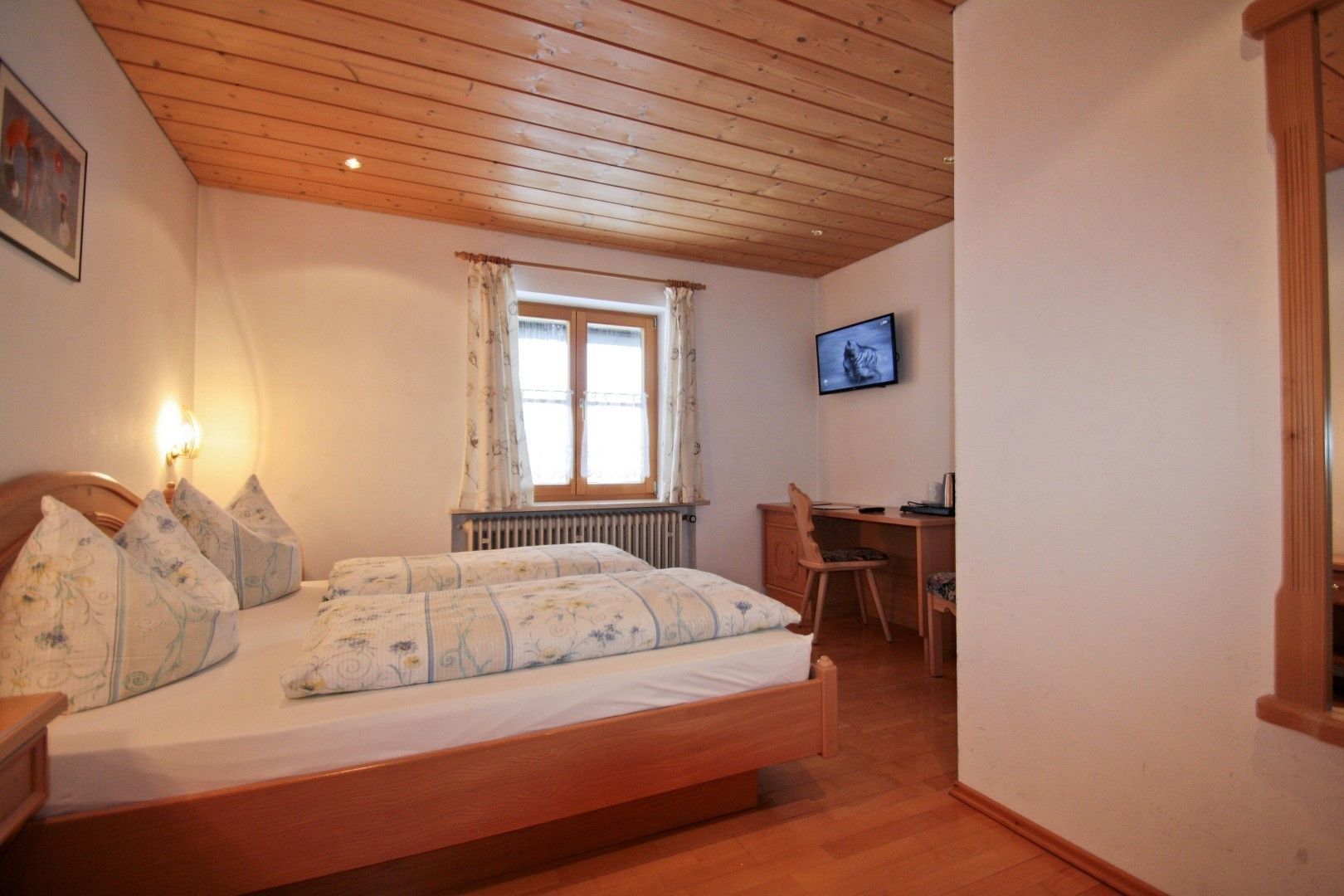 Un dormitorio con techo de madera, una cama con ropa de cama floral, un escritorio con silla y un televisor en la pared.