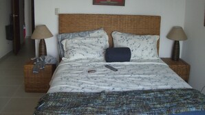 A bed with a woven headboard, two bedside tables with lamps, a remote control, and a framed floral artwork on the wall. - Juan Dolio Apartment (Guayacanes)