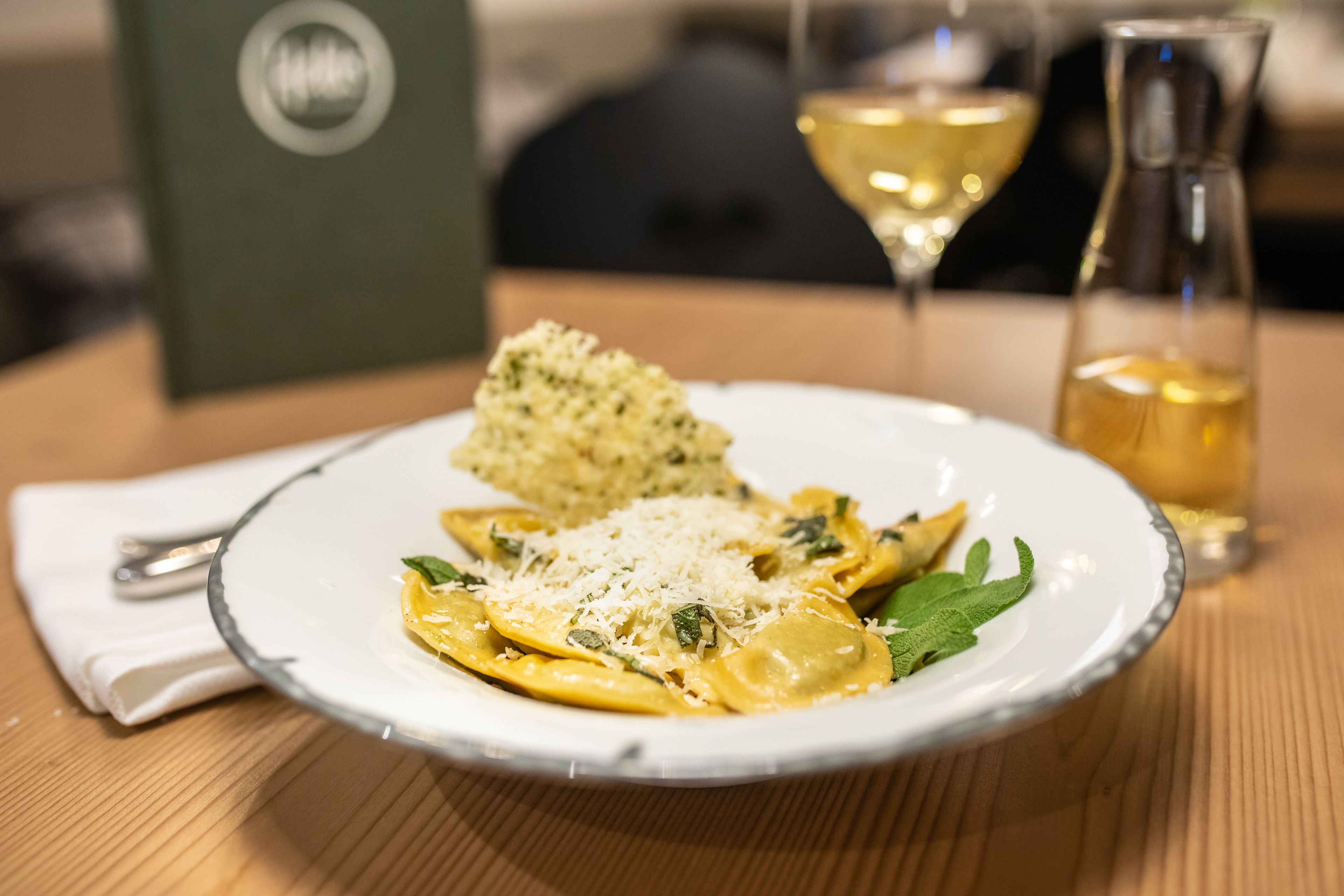 A plate of ravioli with cheese and mint leaves, a glass of white wine, and a bottle of olive oil on a wooden table.