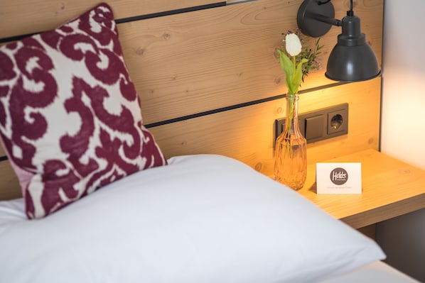 Una cama bien hecha con un cojín estampado, una mesita de noche con una lámpara y una tarjeta sobre la mesa.