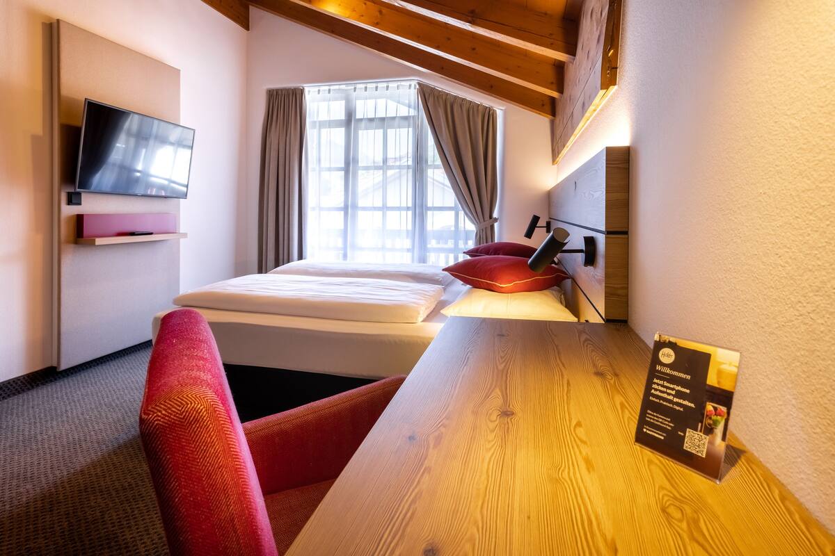 A hotel room with a bed, a TV, a wooden table, a red chair, and a window with curtains.