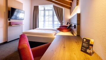 A hotel room with a bed, a TV, a wooden table, a red chair, and a window with curtains.