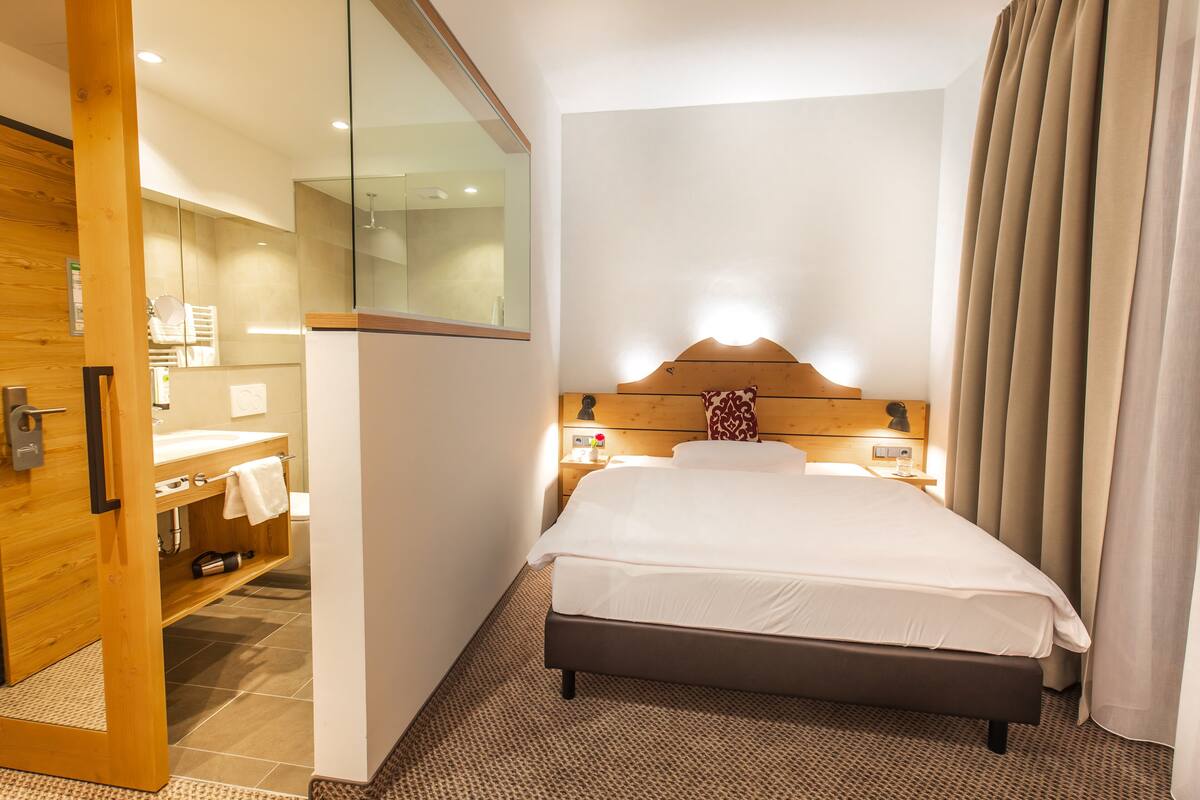 A hotel room with a bed, a wooden headboard, a nightstand with a lamp, a mirror, and a bathroom with a sink and towel rack.