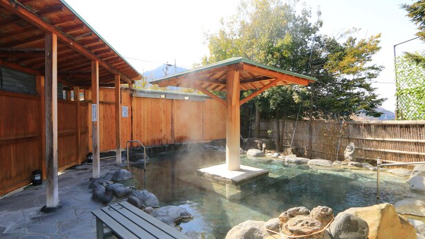 Un balneario de madera tradicional con suelo de piedra, un banco de madera y un área de aguas termales con rocas y una estructura de madera en lo alto.