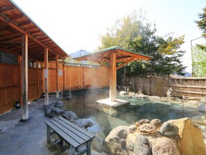 A traditional wooden bathhouse with a stone floor, a wooden bench, and a hot spring area with rocks and a wooden structure overhead.
