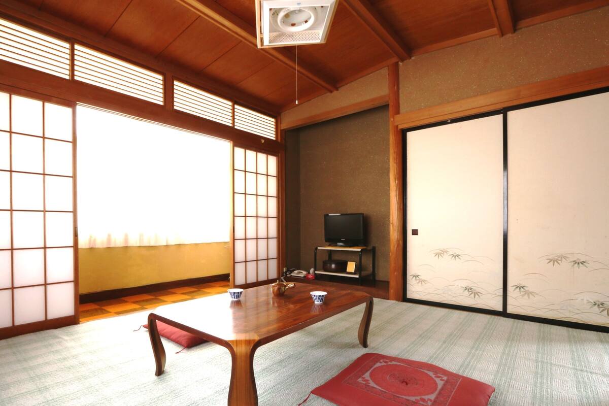 Una habitación japonesa tradicional con suelo de tatami, una mesa de madera baja, un televisor sobre un mueble y puertas correderas con diseño de bambú.
