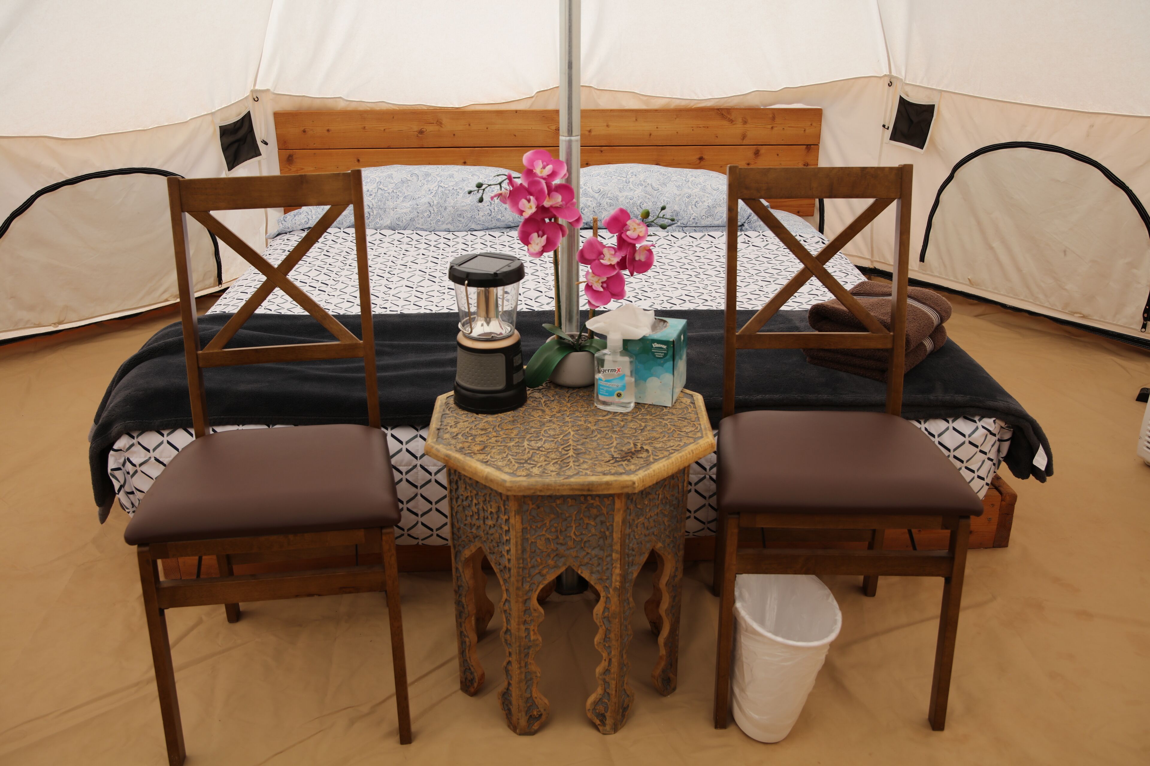 Een tent met een bed, twee stoelen, een kleine tafel met een lamp en een prullenbak.