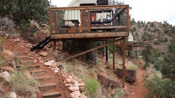 A rustic wooden cabin with a staircase leading up to it, surrounded by rocky terrain and sparse vegetation.