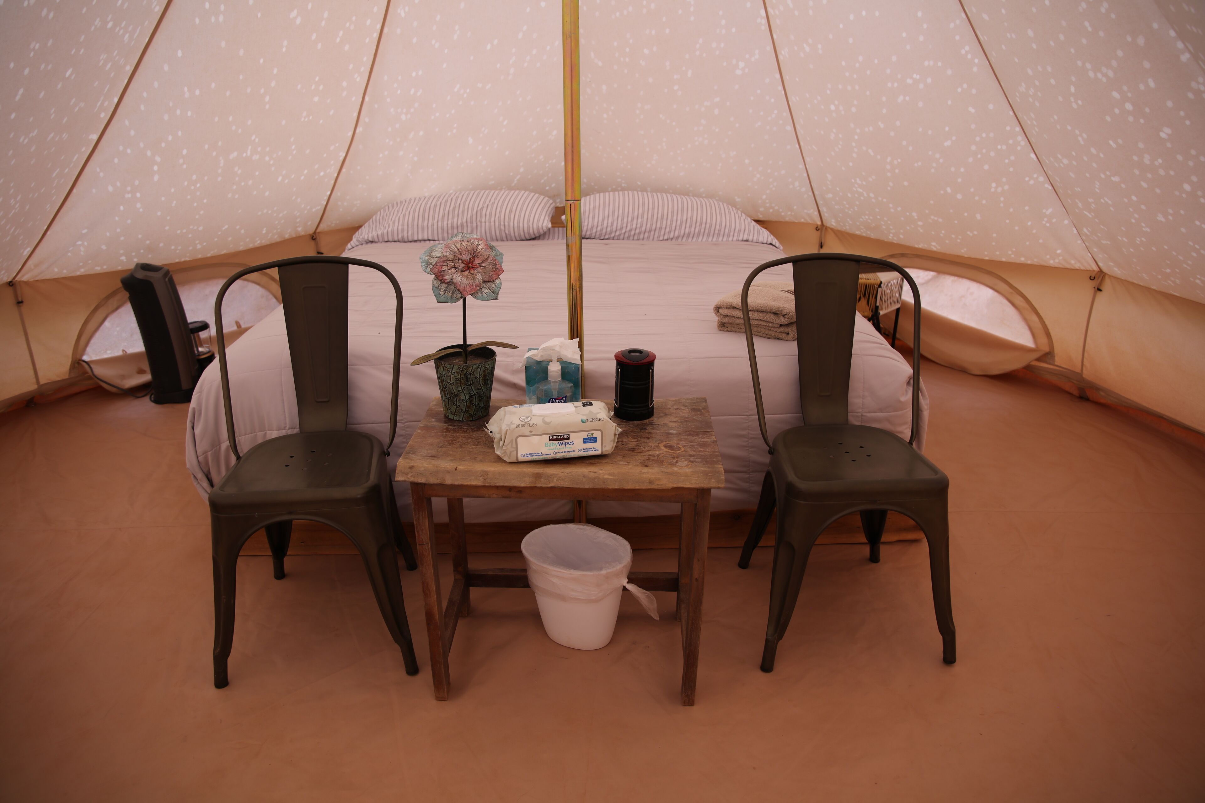 A tent with a bed, two chairs, a table, a bucket, and a small plant.