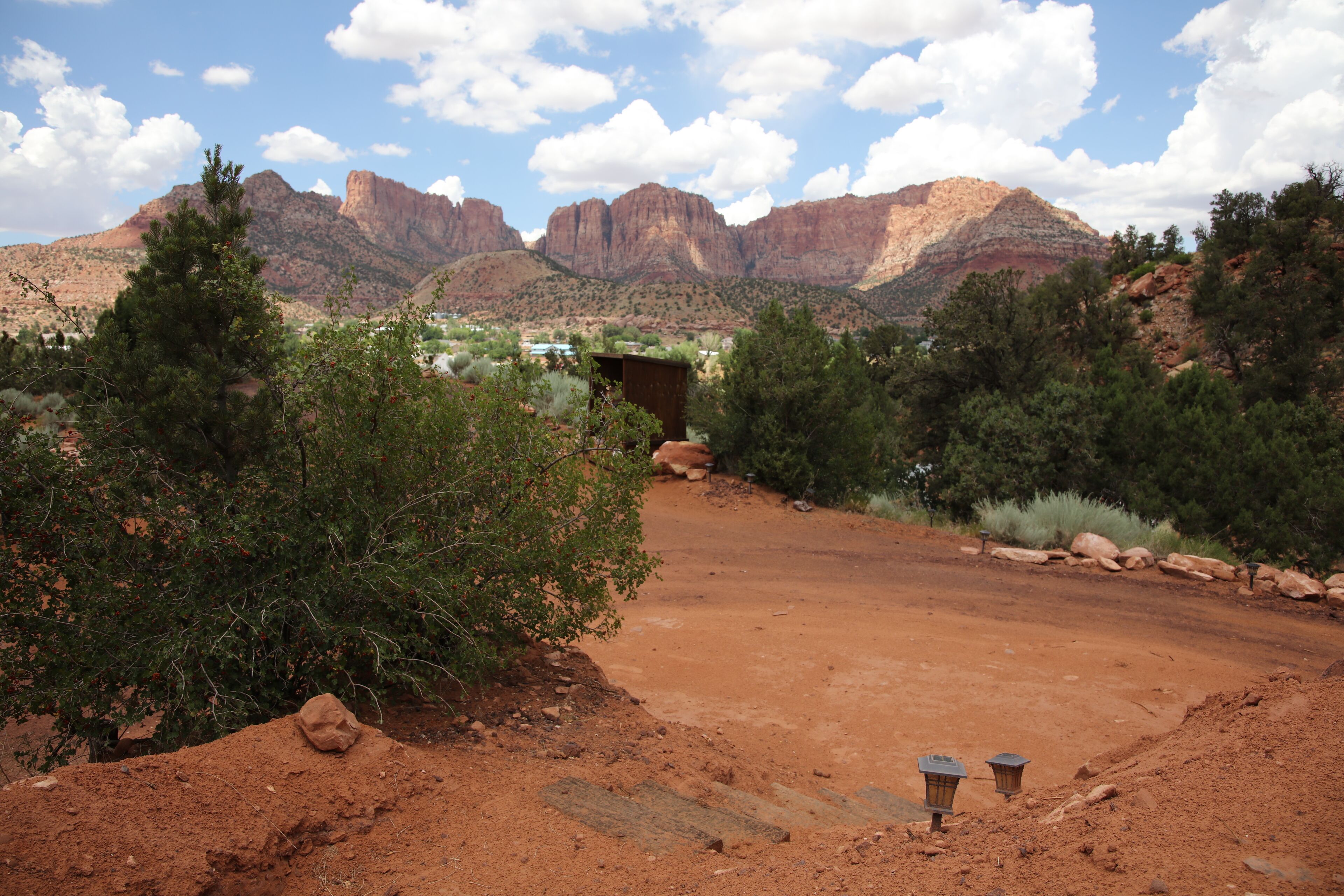 A desert landscape with a dirt path leading to a small building, surrounded by trees and mountains in the background.