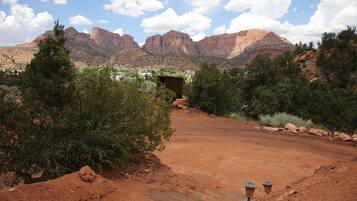A desert landscape with a dirt path leading to a small building, surrounded by trees and mountains in the background.