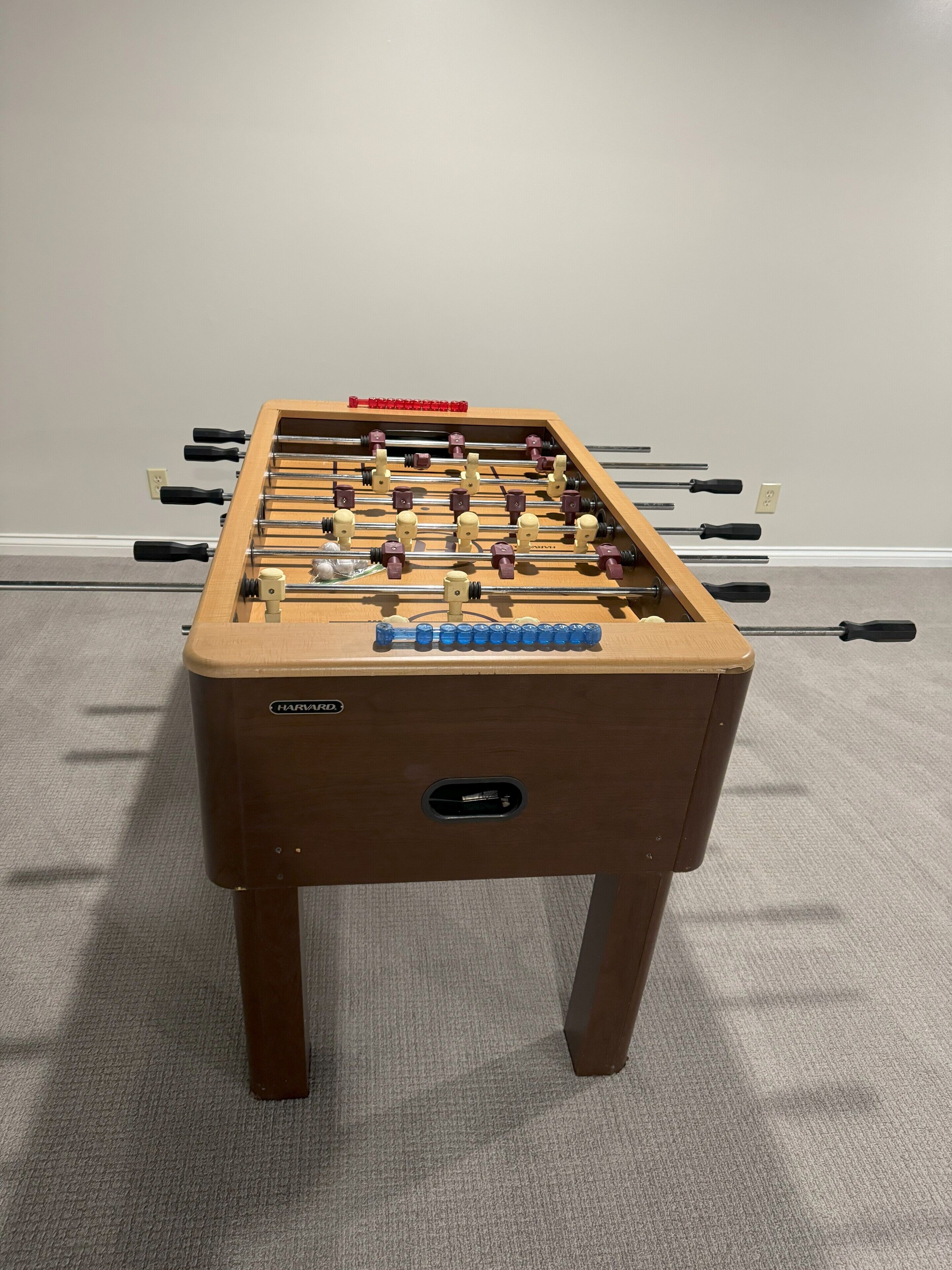 Una mesa de fútbol de mesa con base de madera y panel de control, frente a una pared lisa con suelo alfombrado.