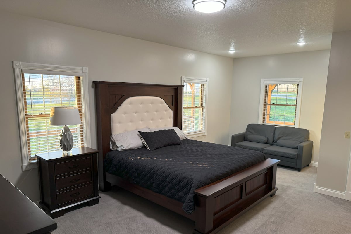 A bedroom with a large bed, a nightstand, a lamp, a gray sofa, and windows with blinds.