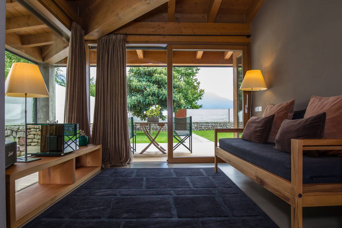 Quarto aconchegante com um banco de madeira, uma mesa com luminária e vista para um jardim e montanhas.