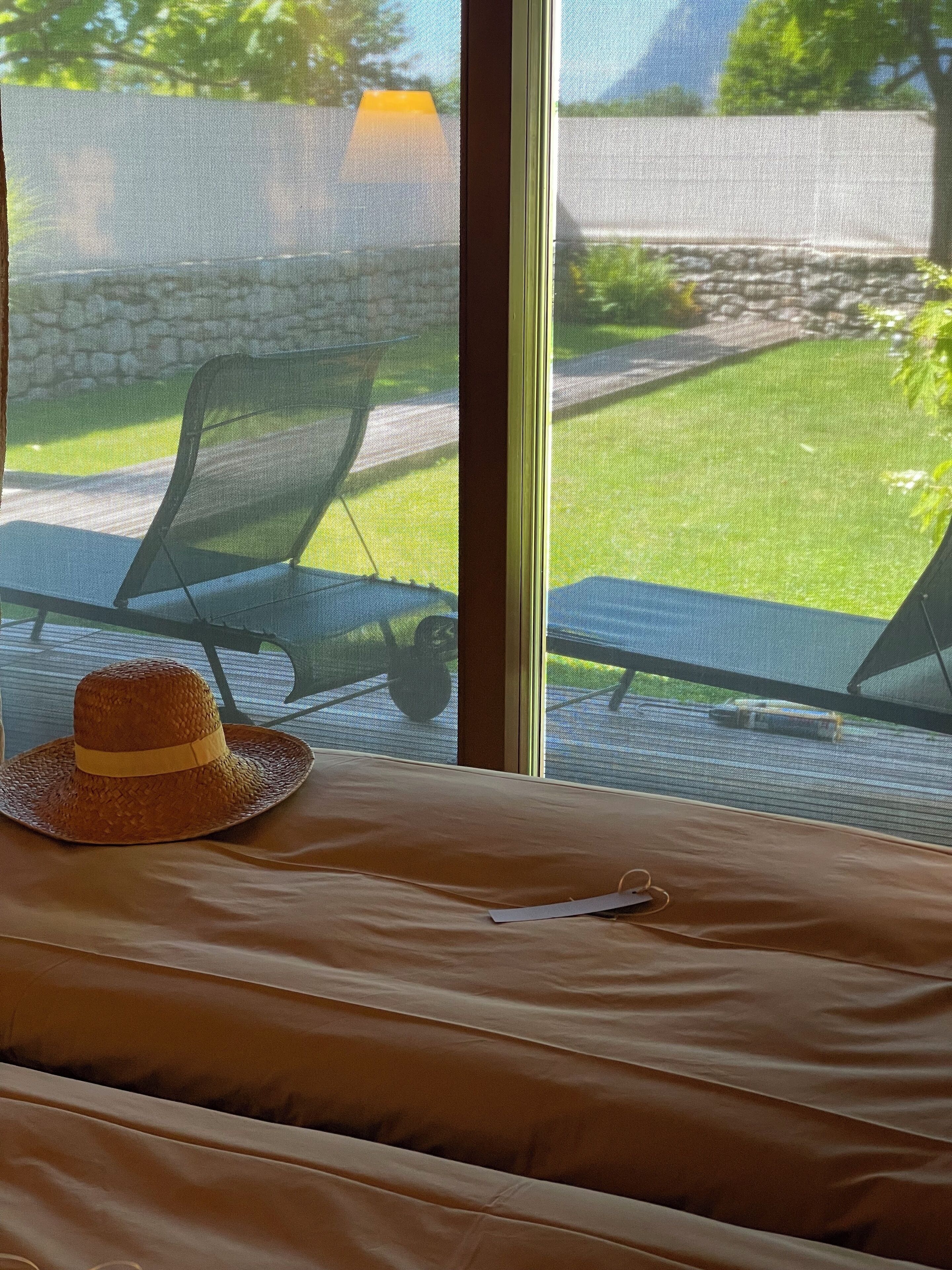 Ein Bett mit einem Strohhut, ein Fenster mit Blick auf eine Rasenfläche und Stühle.