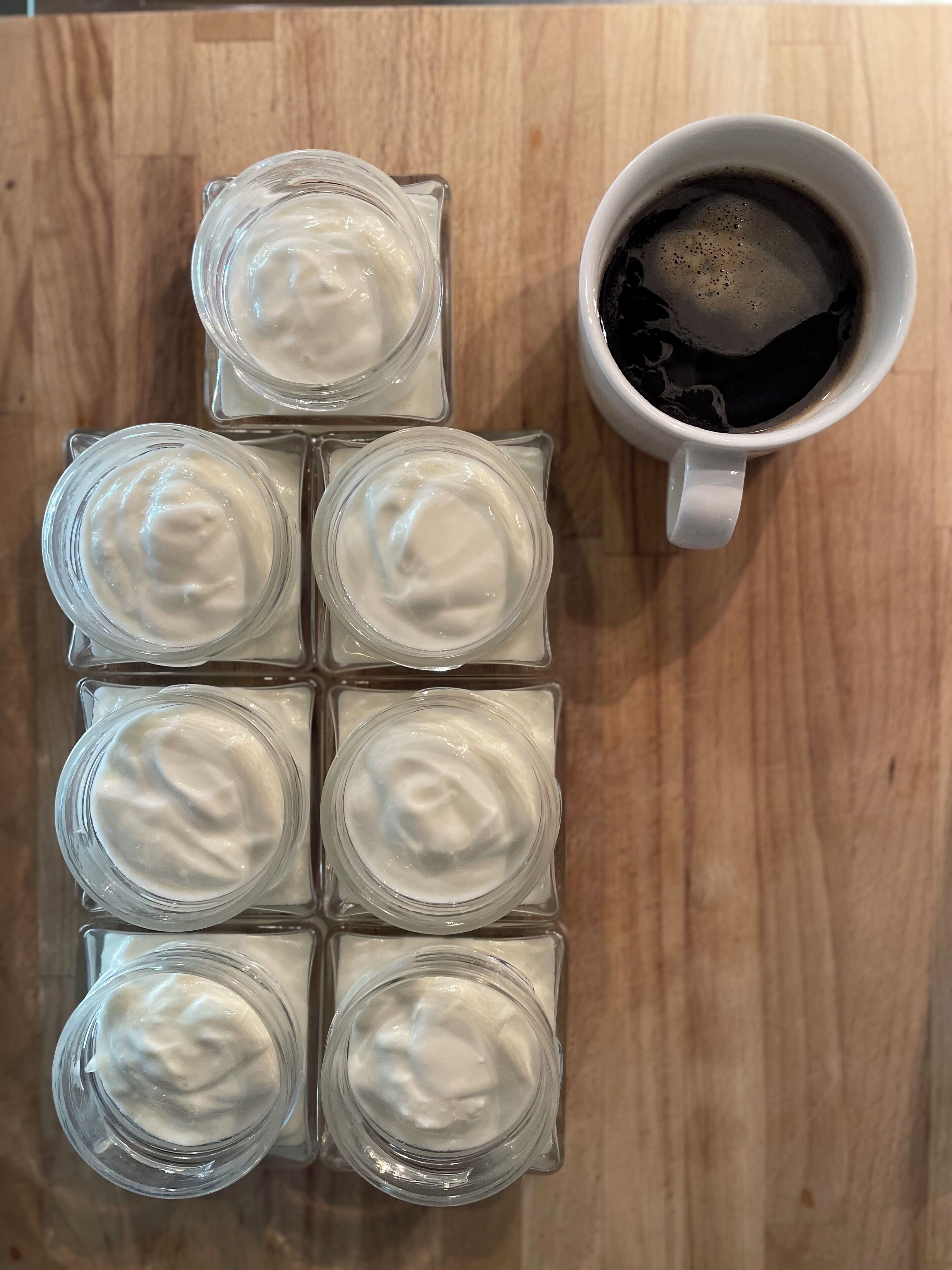 Sechs klare Glasbehälter mit weißer Substanz und eine weiße Tasse mit schwarzer Flüssigkeit auf einer Holztischfläche.