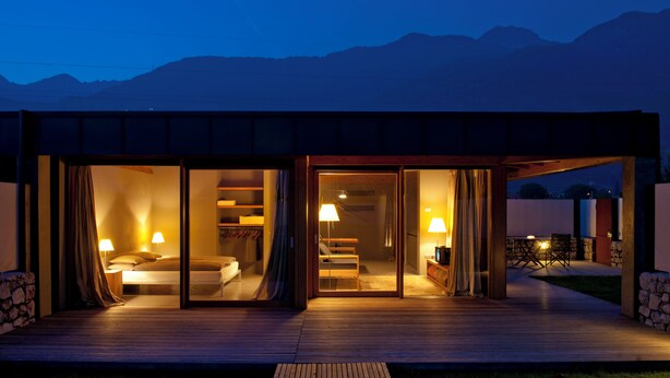 A modern house with a wooden deck, large sliding glass doors leading to a bedroom with a bed and two lamps, and a staircase. - VIVERE suites and rooms (Arco)