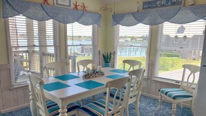 A dining area with a white table and chairs, blue placemats, and a view of the outdoors through large windows. - Two If By Sea I and II: Larger home with fabulous views of Silver Lake (Ocracoke)