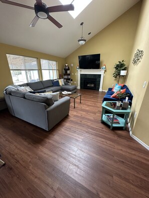 A living room with a sofa, a coffee table, a fireplace, a TV, and a ceiling fan. - Oregon Coast Lake front Oasis (Warrenton)