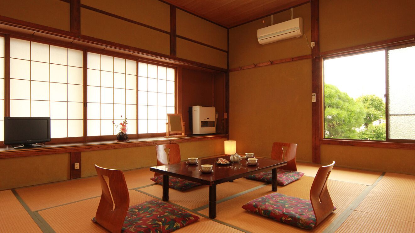 Habitació japonesa tradicional amb fons de tatami, una taula baixa amb coixins, una televisió i una finestra amb vista a vegetació.