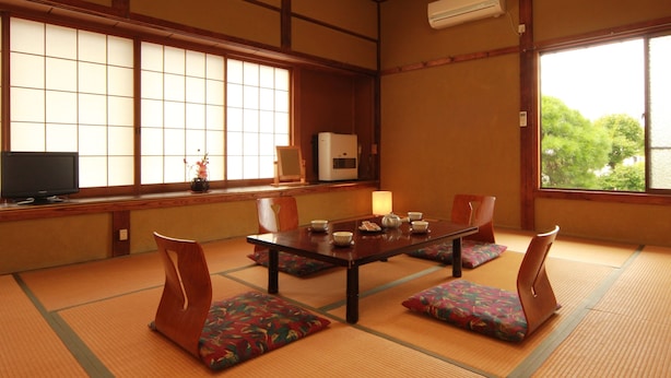 Une pièce de style japonais traditionnel, avec un sol recouvert de tatamis, une table basse avec des coussins, un téléviseur et une fenêtre donnant sur un paysage verdoyant.