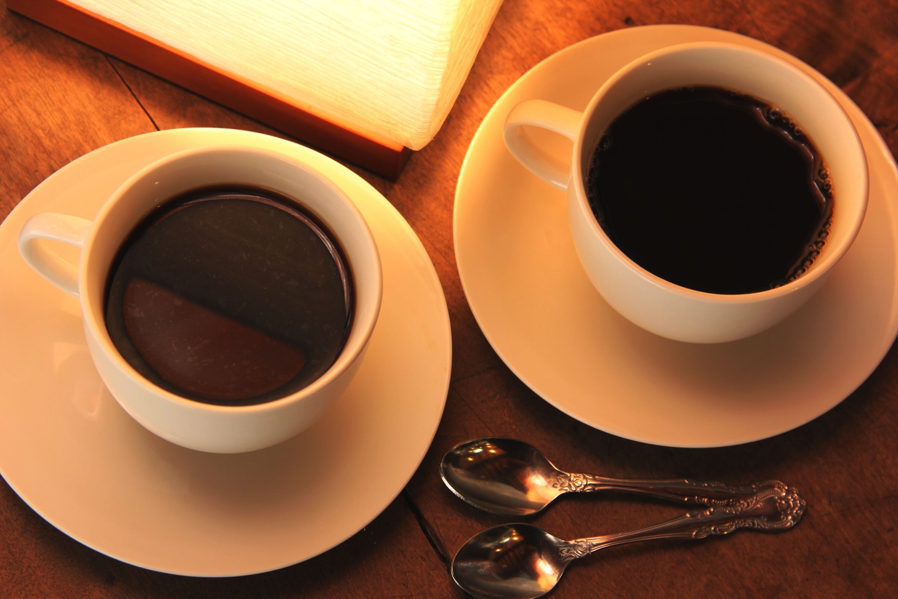 Zwei weiße Kaffeetassen mit Untertassen und Löffeln auf einem Holztisch.