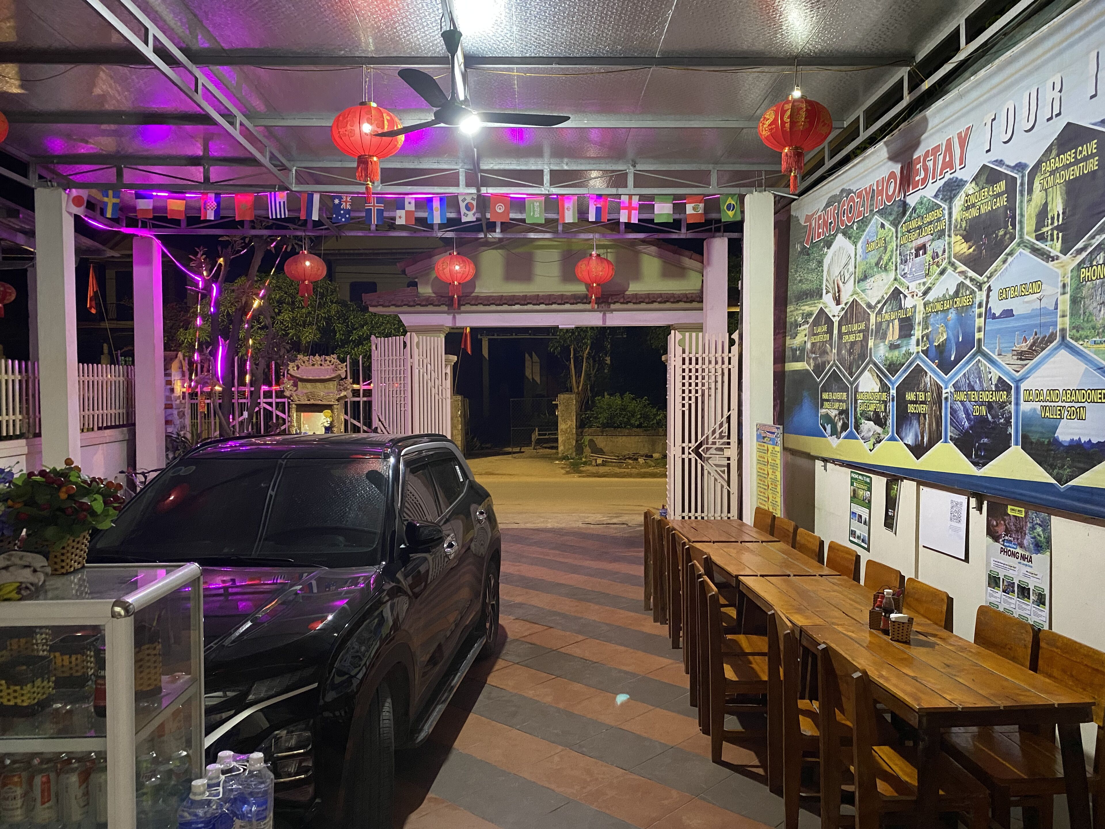 Un restaurante con un coche estacionado afuera, mesas y sillas en el interior, y una pared decorada con varios carteles.