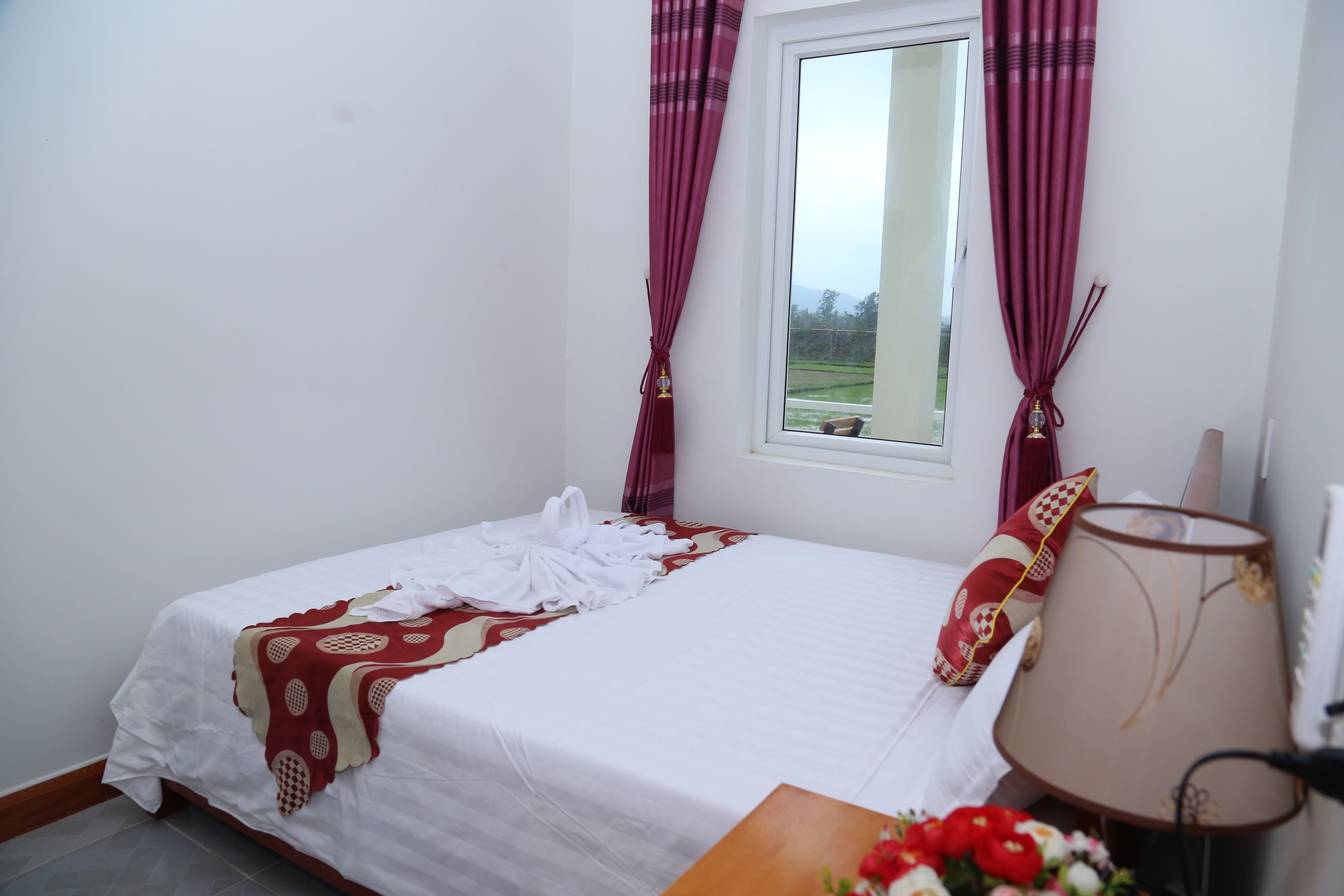 A neatly made bed with a white and red patterned blanket, a wooden nightstand with a lamp, and a window with curtains.