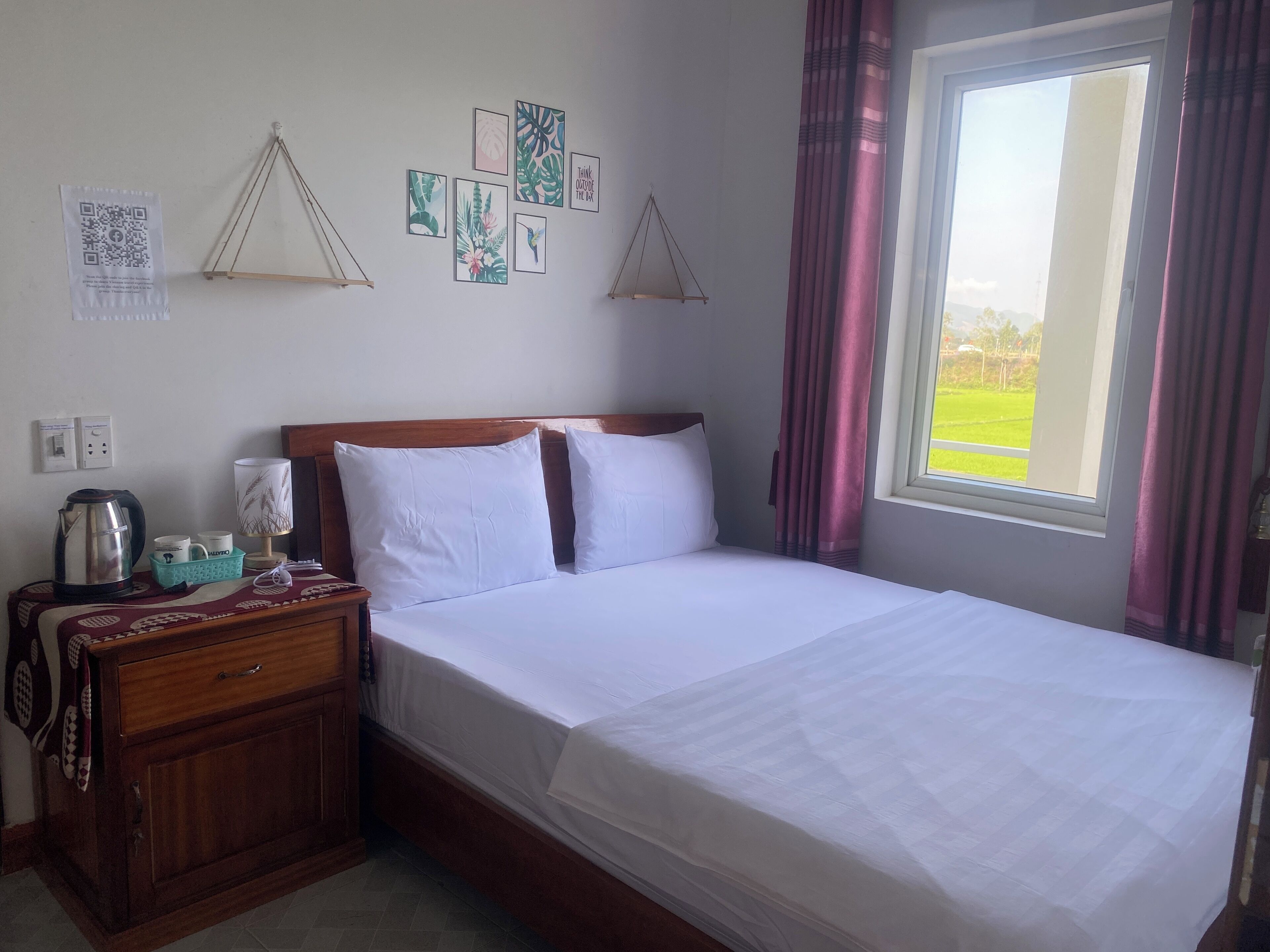 A hotel room with a bed, a nightstand with a kettle, cups, and a lamp. There are wall decorations and a window with curtains.
