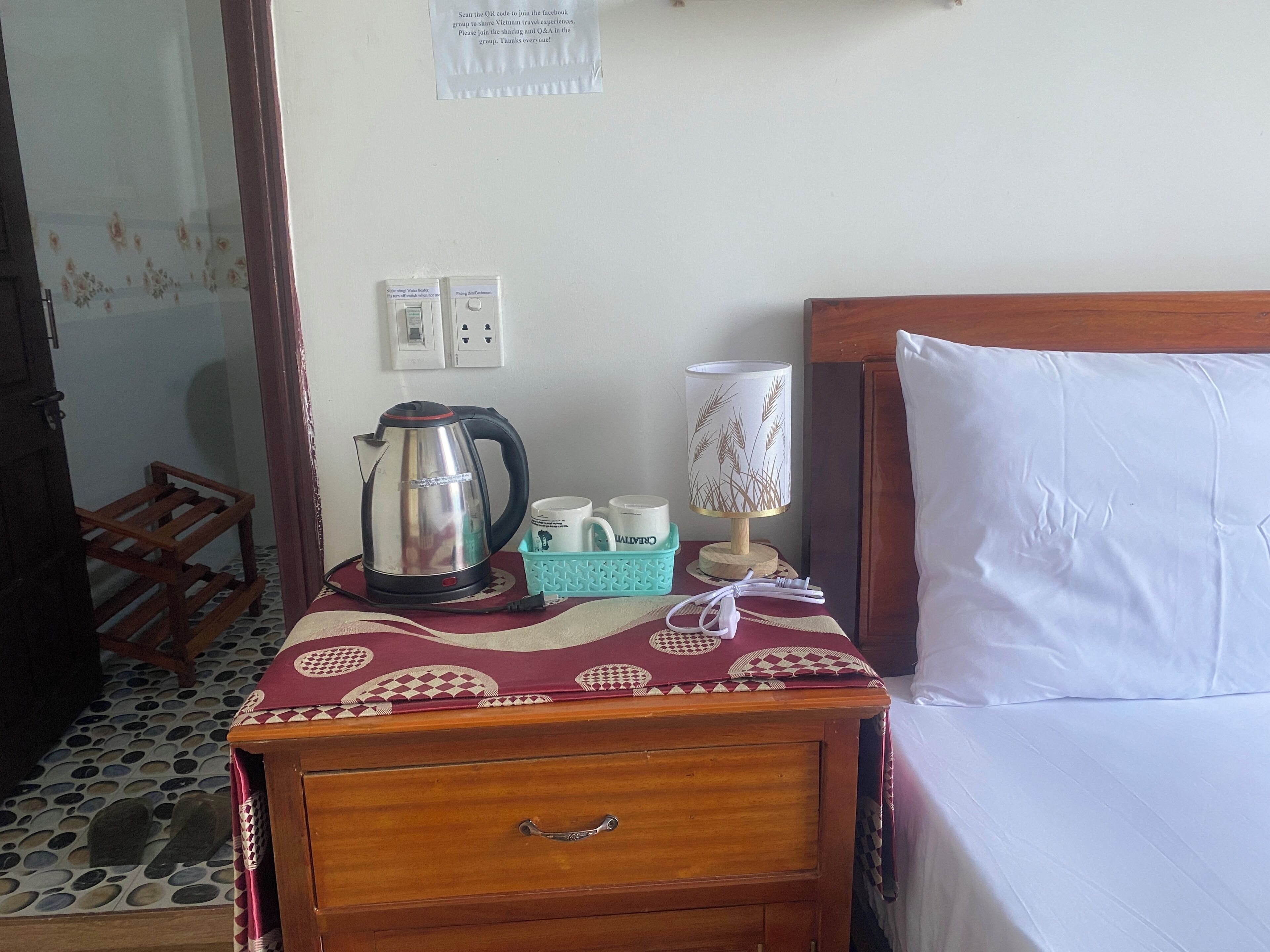 A hotel room with a wooden nightstand, a bed with white sheets, a kettle, a lamp, and a small table with a tray holding cups and a container.