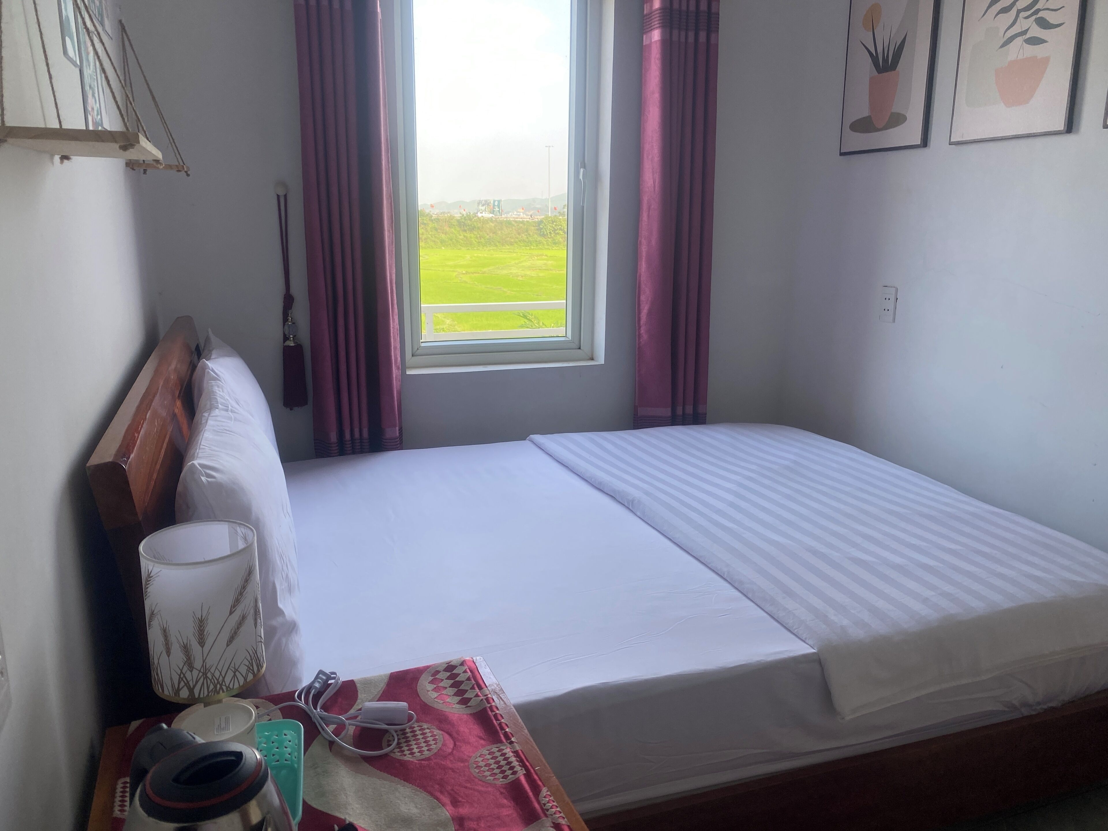 Una cama individual con cabecera de madera, una mesita de noche con lámpara, una regleta eléctrica y una ventana con cortinas.