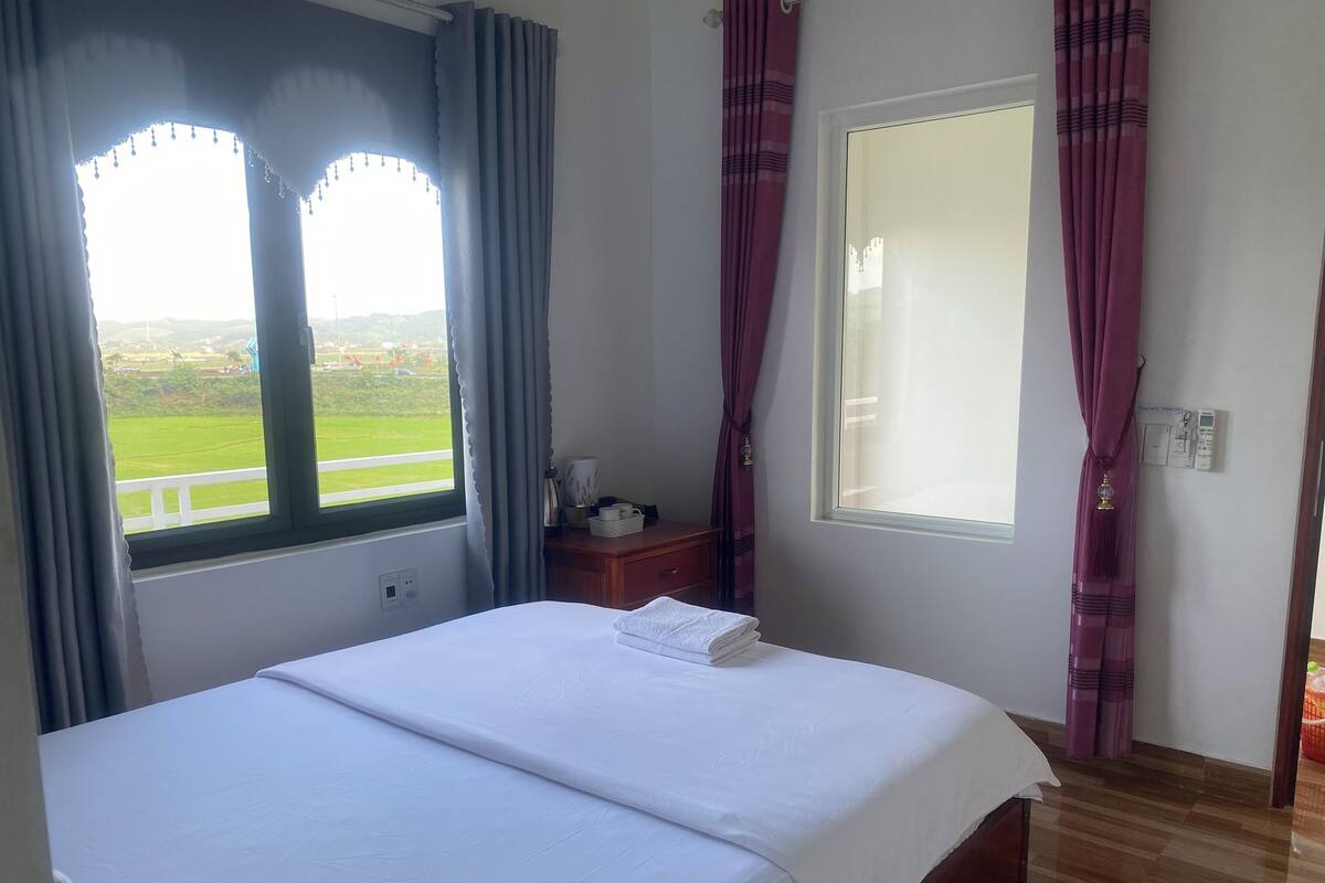 A hotel room with a bed, a nightstand, a window with curtains, and a view of a green landscape.