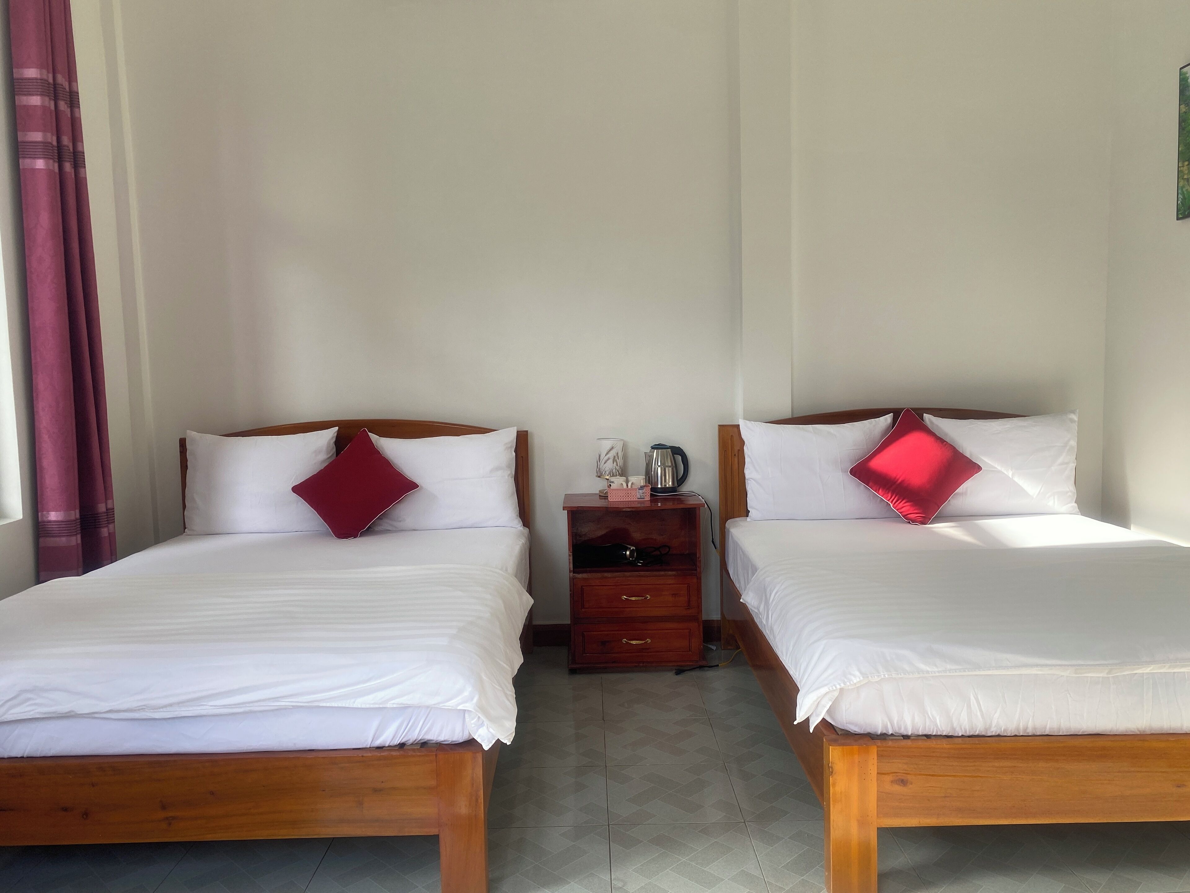 A room with two wooden beds, each with white bedding and a red pillow. There is a wooden nightstand between the beds with a kettle and a cup on it.