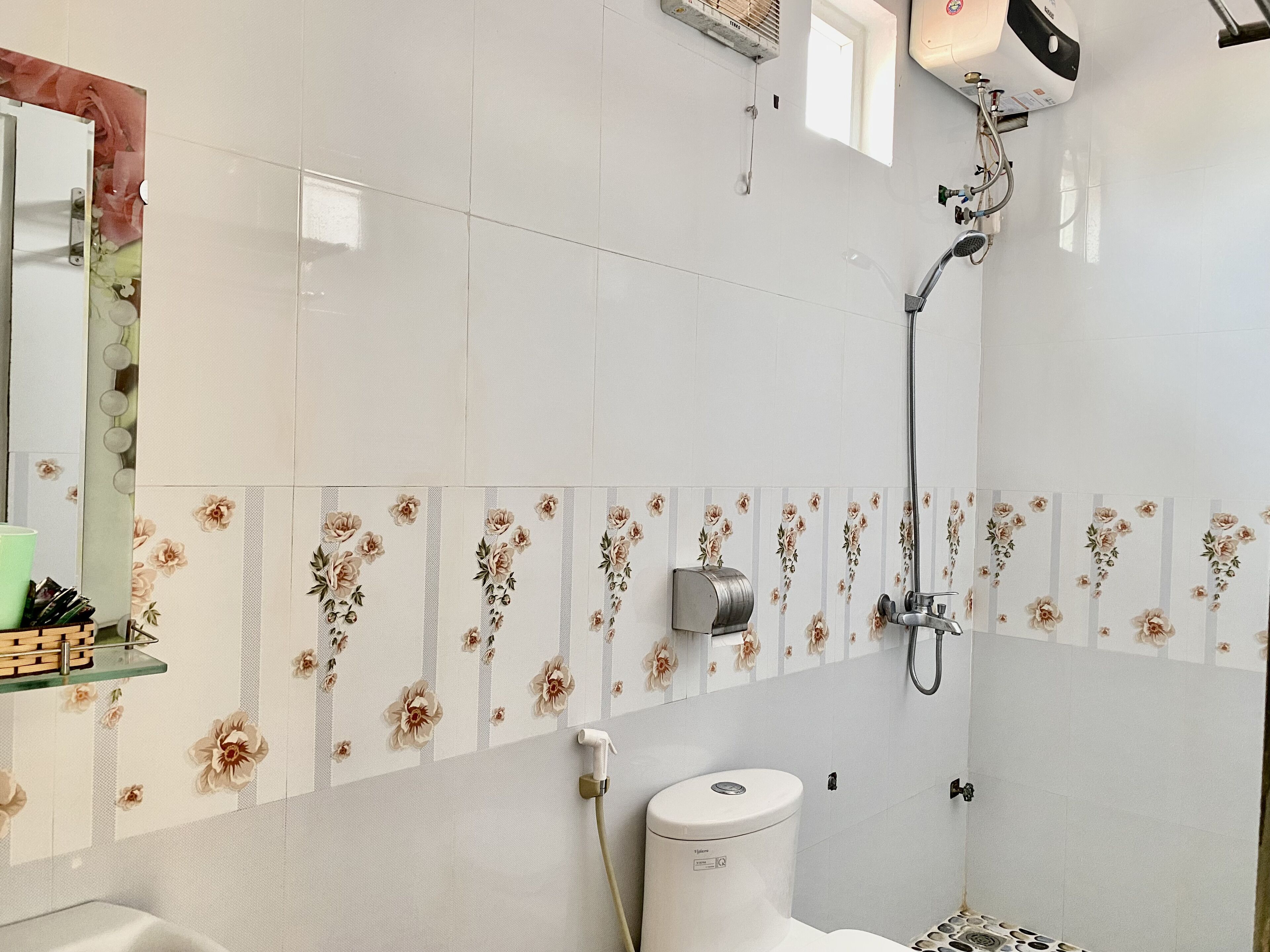 A bathroom with white tiles, a shower, a toilet, and a shelf with toiletries.