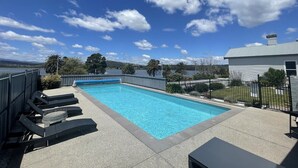 A rectangular swimming pool with clear blue water, surrounded by a concrete deck. There are lounge chairs along the poolside, and a fence enclosing the area.