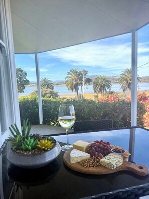 A table with a cheese platter, grapes, nuts, and a glass of white wine, overlooking a scenic view of a lake and palm trees.