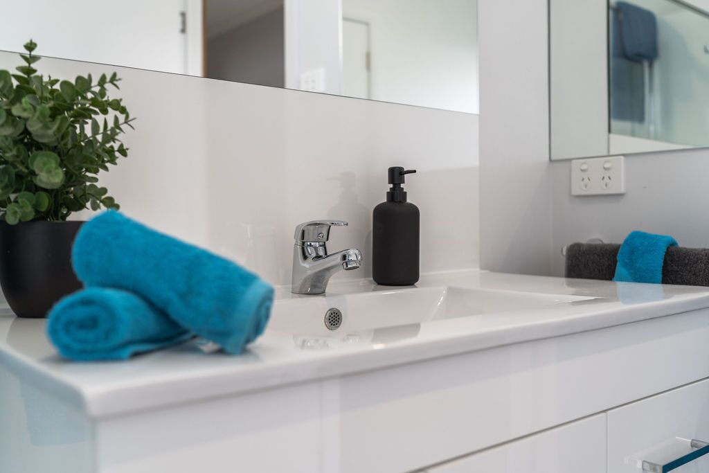 Ein modernes Badezimmer mit einem weißen Waschbecken, einem schwarzen Seifenspender, einem blauen Handtuch und einer Topfpflanze.