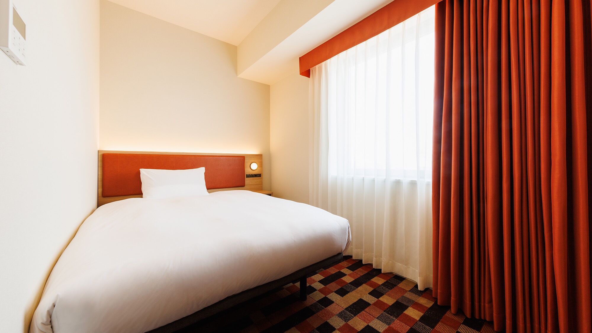 A hotel room with a bed, a nightstand, a wall-mounted lamp, a window with curtains, and a patterned carpet.
