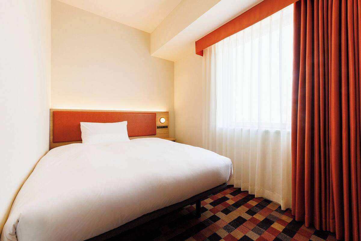 A hotel room with a bed, a nightstand, a wall-mounted lamp, a window with sheer curtains, and a patterned carpet.