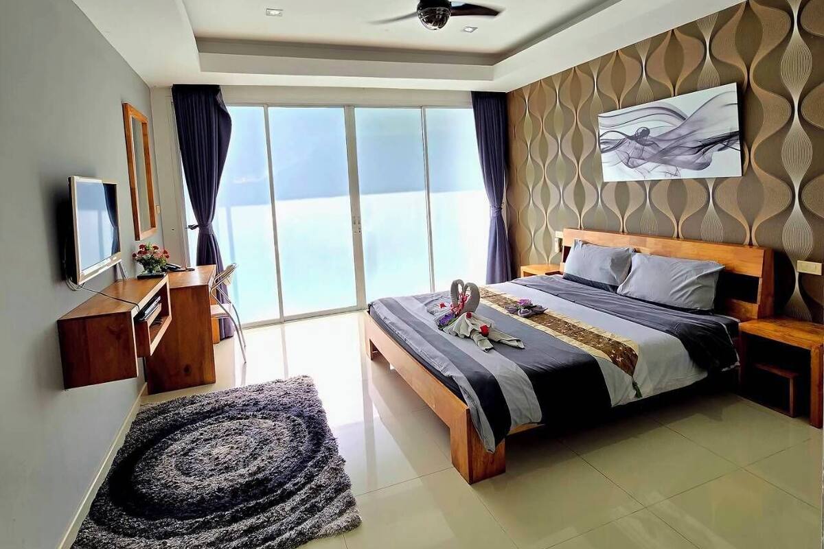 Une chambre d’hôtel moderne dotée d’un grand lit, d’un ventilateur de plafond, d’une télévision et offrant une vue sur la mer grâce à de grandes fenêtres du sol au plafond.