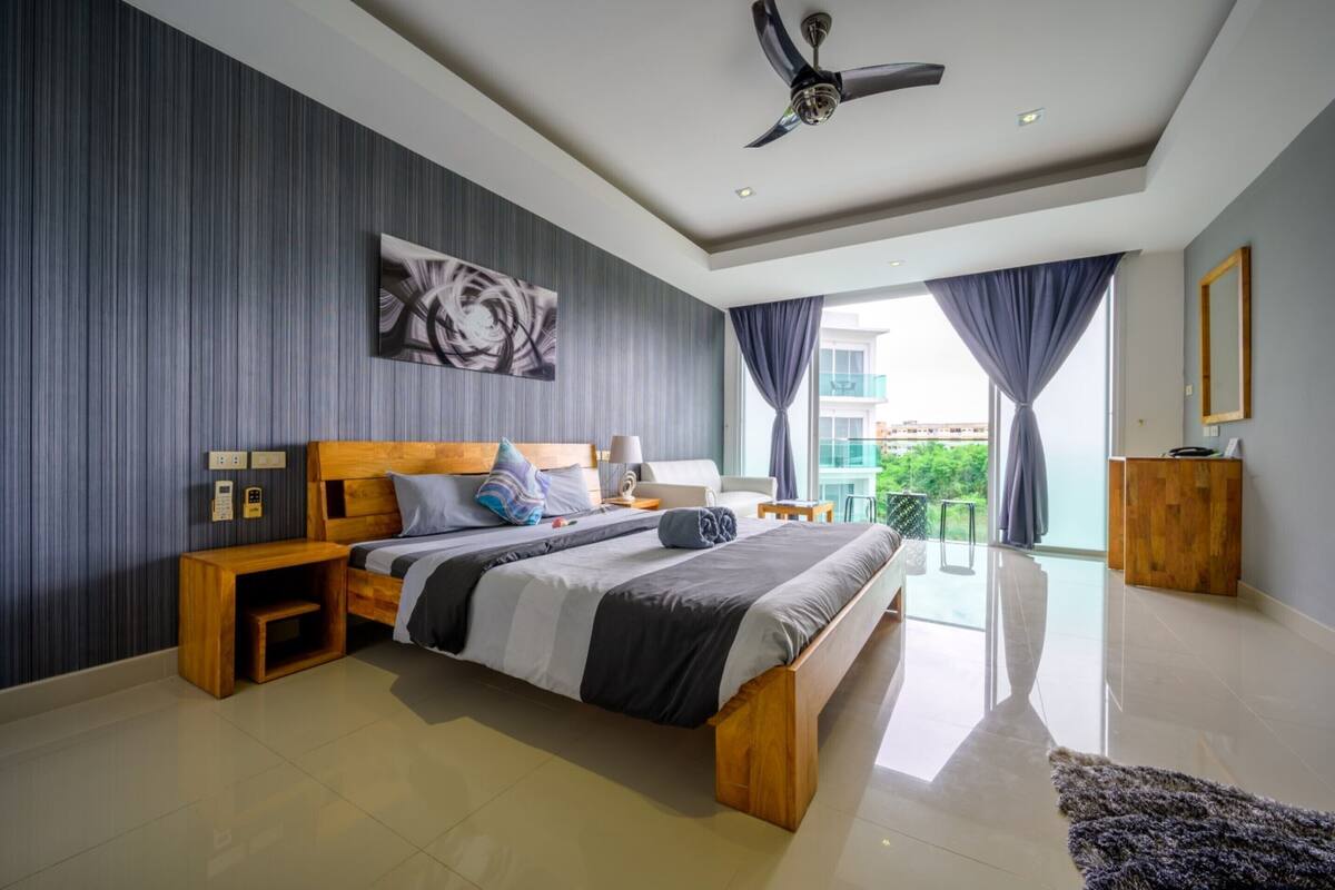 Une chambre moderne avec un grand lit, une tête de lit en bois, des tables de chevet, un ventilateur de plafond et une vue sur l’extérieur grâce à de grandes fenêtres.