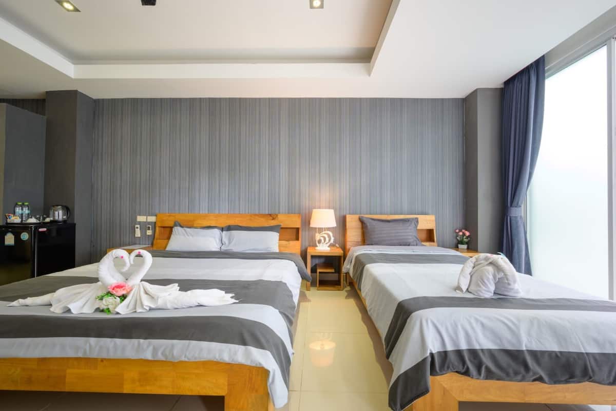 Une chambre d’hôtel avec deux lits, une tête de lit en bois, une table de chevet avec une lampe, une petite table avec des fleurs et une grande fenêtre avec des rideaux.