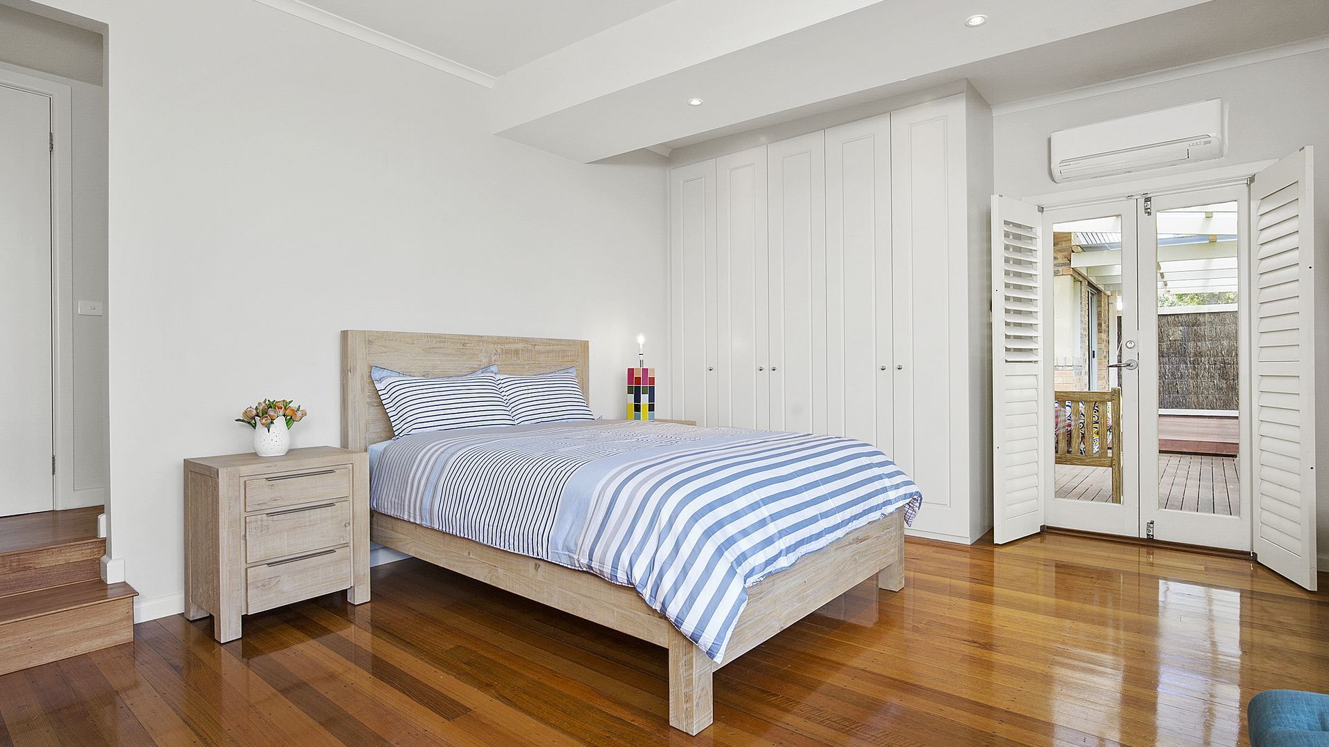 A bedroom with a wooden bed, white walls, and a wooden floor. There is a nightstand with a vase of flowers, a wardrobe, and a door leading to a balcony.