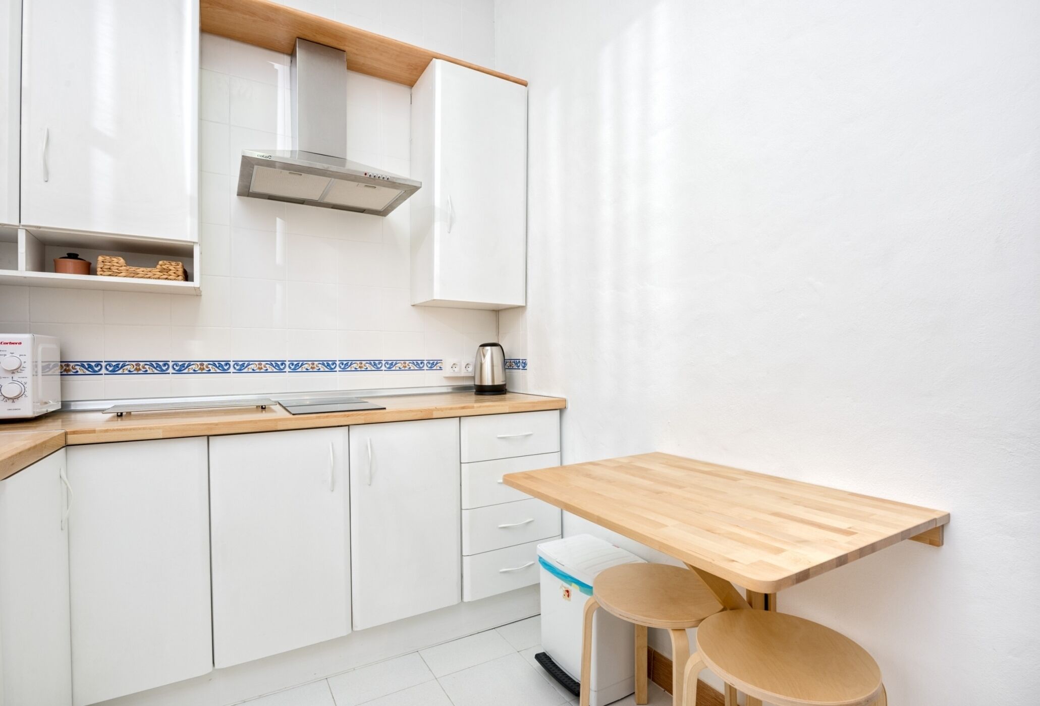 Eine kleine Küche mit weißen Schränken, einer Holz Arbeitsplatte, einem Tisch und einem Wasserkocher.