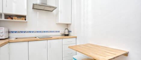 A compact kitchen with white cabinets, a wooden countertop, a small table, and a kettle.