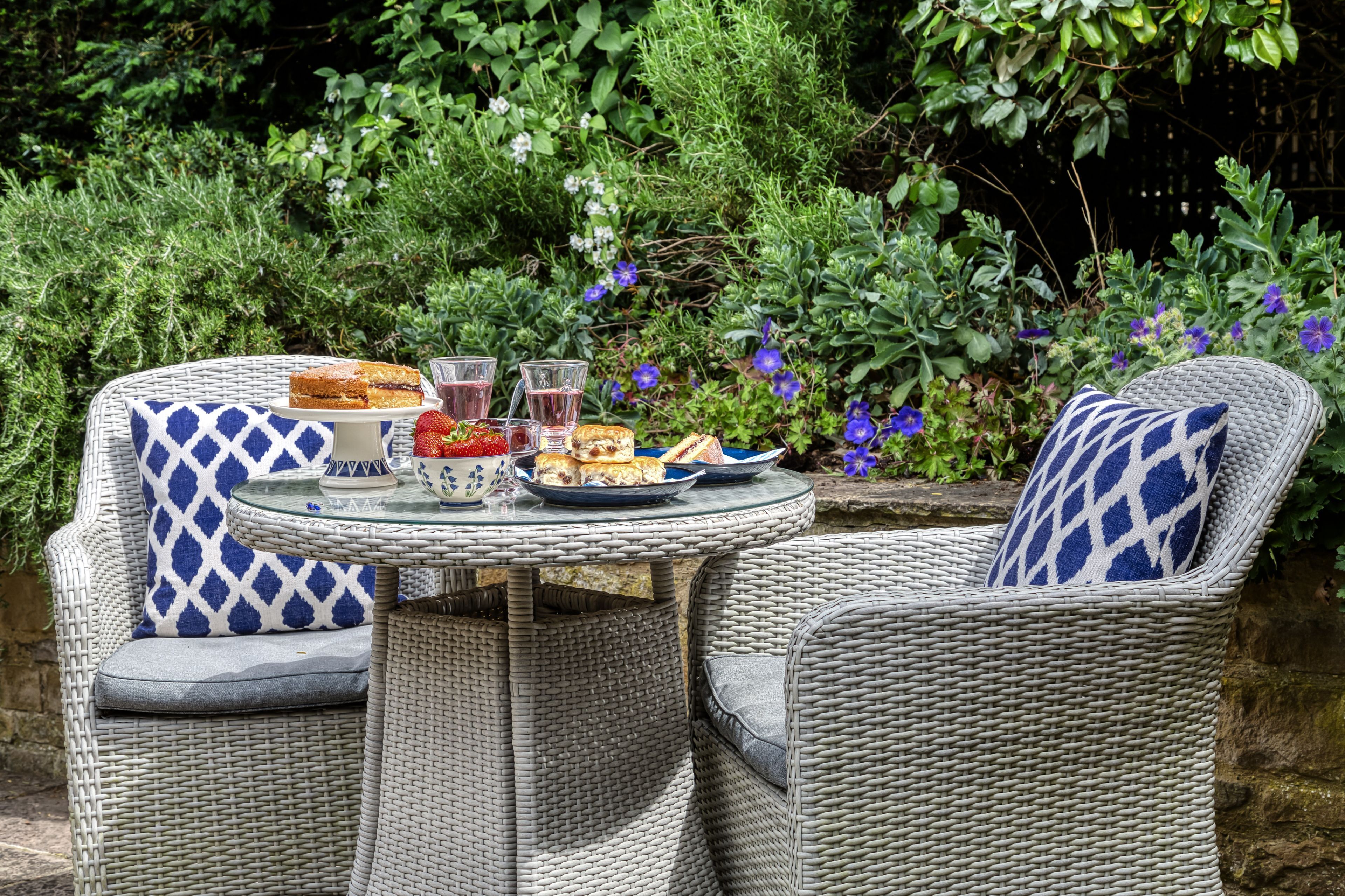 A wicker outdoor dining set with a glass table, two chairs, and a plate of food and drinks.