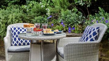 A wicker outdoor dining set with a glass table, two chairs, and a plate of food and drinks.