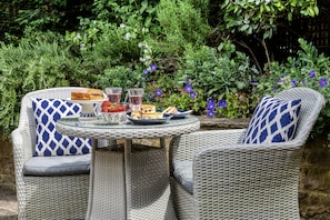A wicker outdoor dining set with a glass table, two chairs, and a plate of food and drinks.